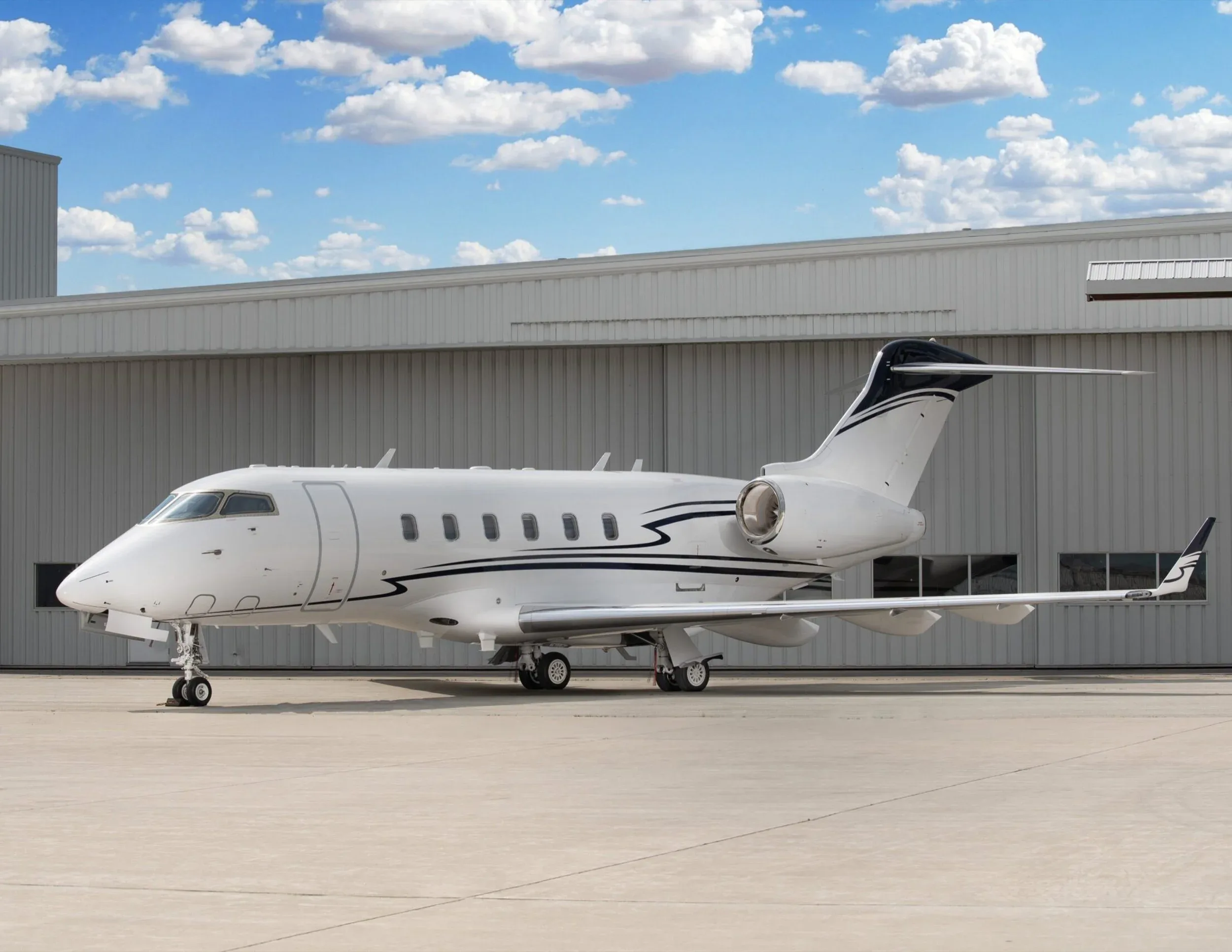 A white business jet with black and blue stripes on the tail and fuselage parked on an tarmac in front of a hangar under a blue sky.