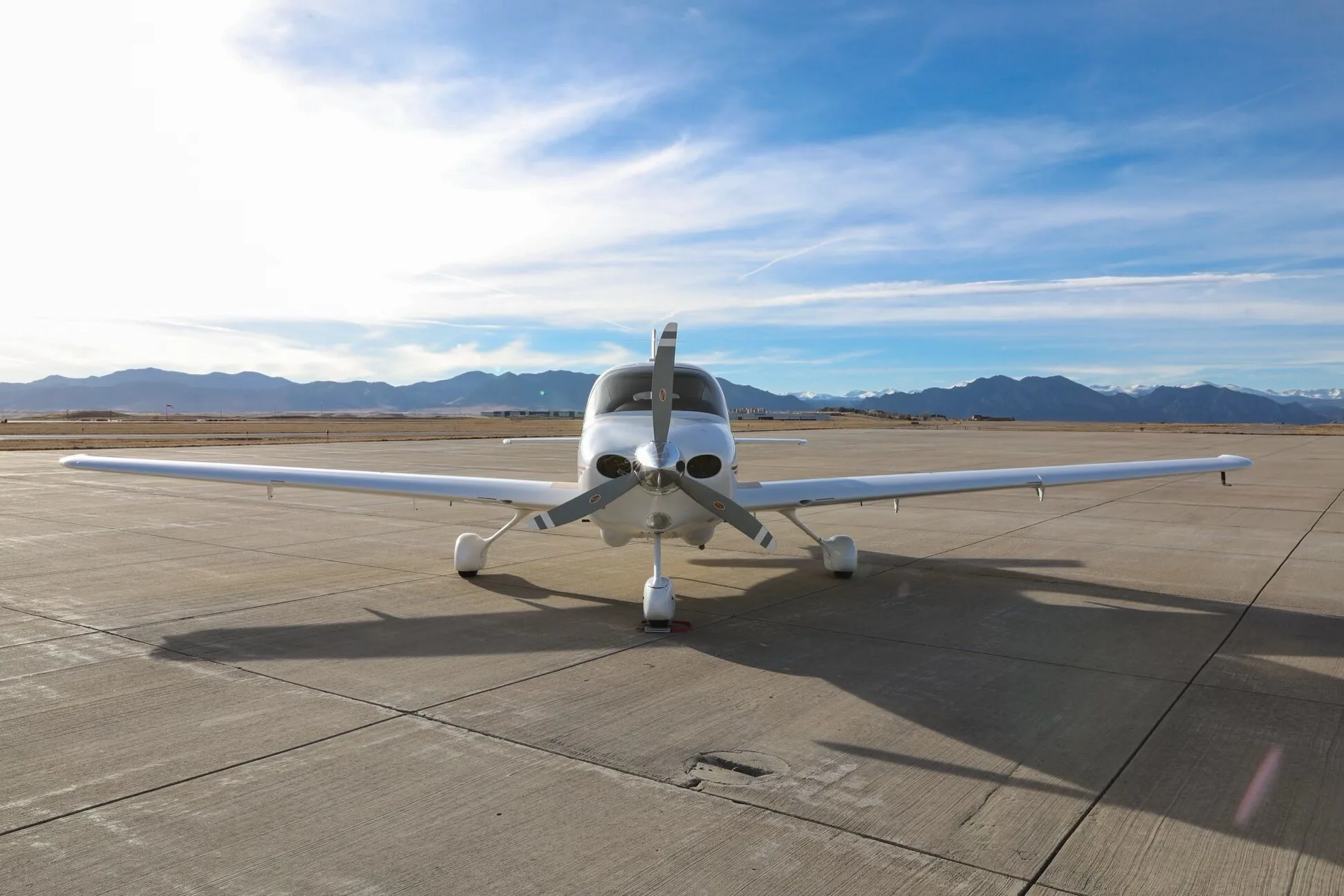Front view of a white Cirrus SR20 aircraft with a three-bladed propeller, parked on an airfield with mountains in the distance.