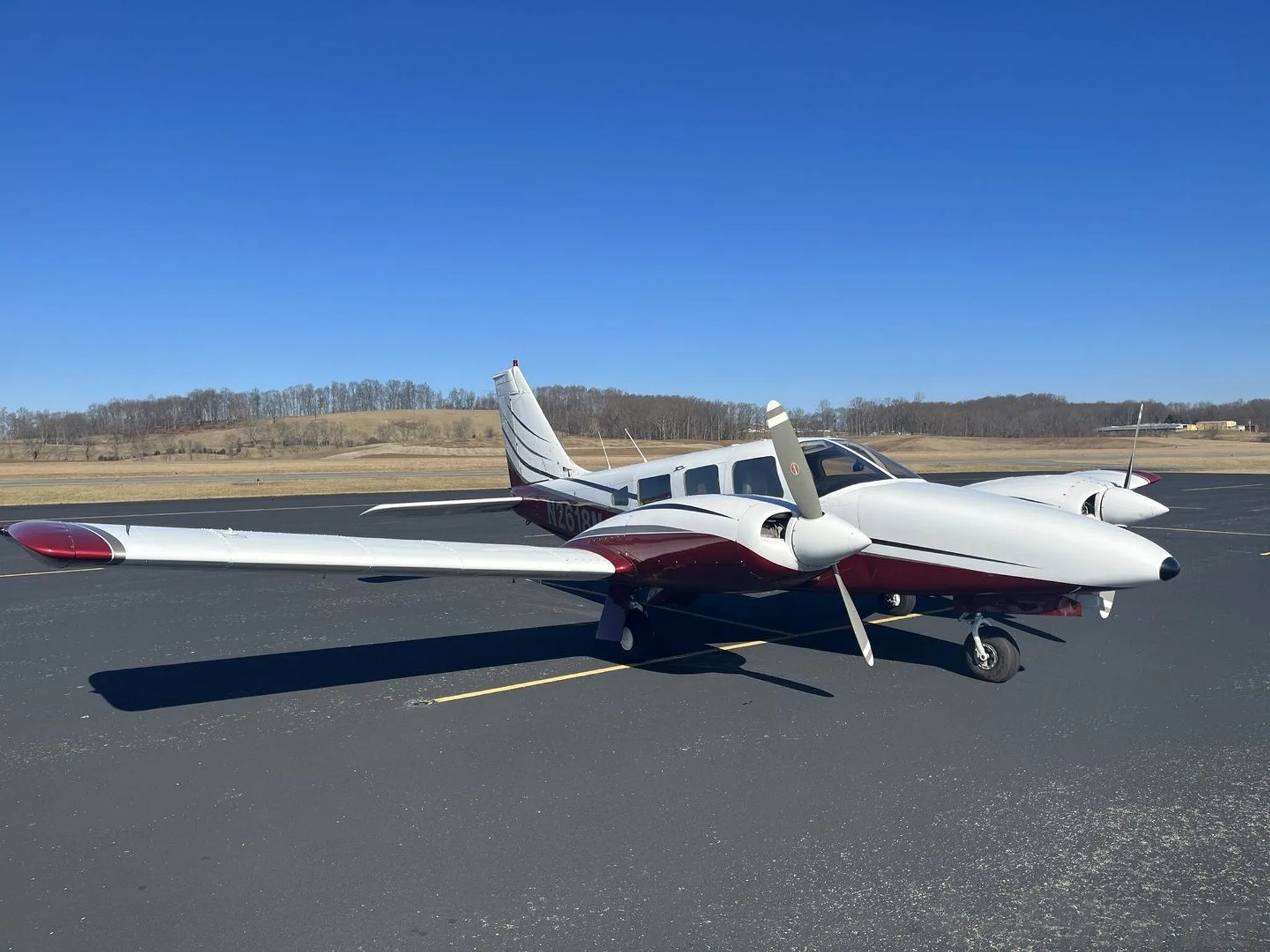 A white and maroon twin-engine Piper aircraft, N2618M, is parked on an asphalt tarmac under a clear blue sky.