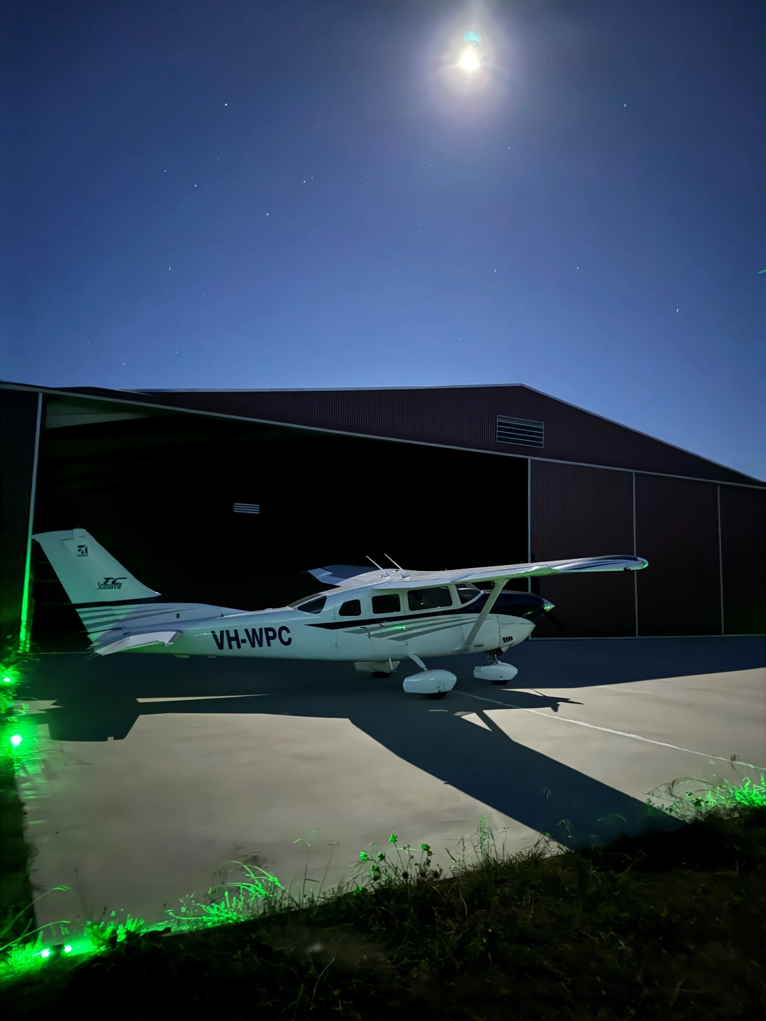 A white Cessna 210 with blue stripes, registration VH-WPC, parked outside a hangar at night under a full moon.