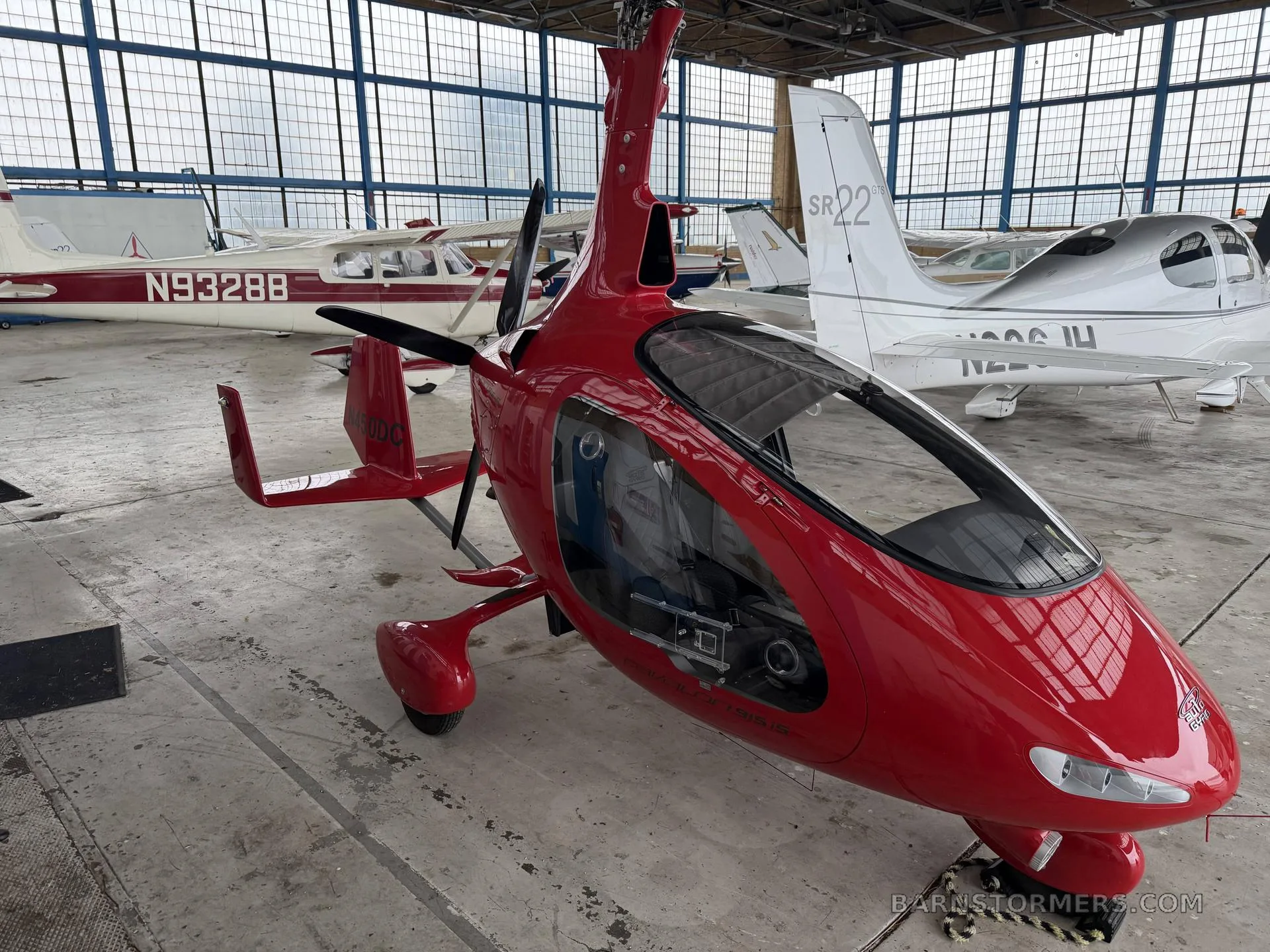 A red AutoGyro Cavalon 915iS autogyro parked in a hangar with other light aircraft.