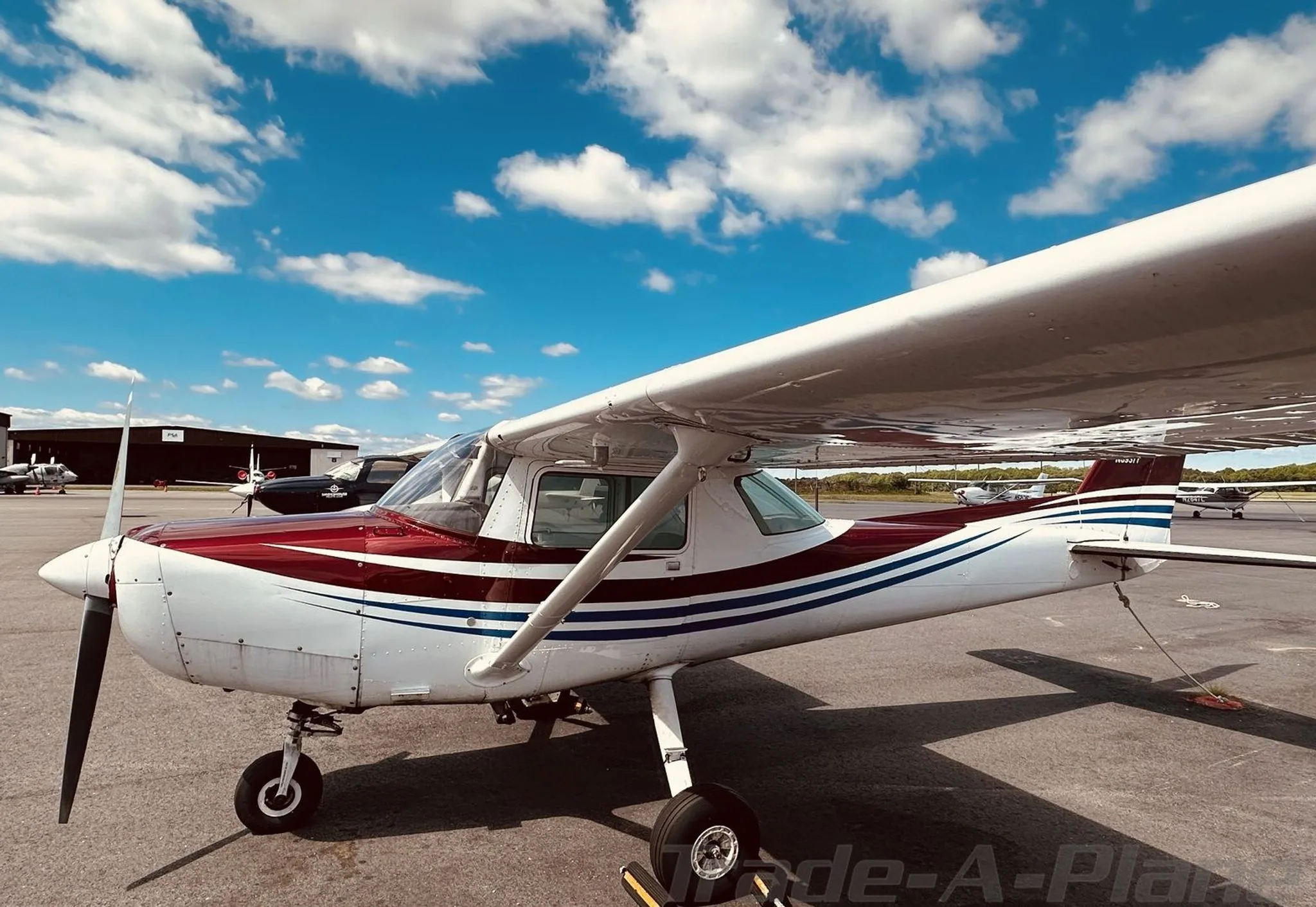A side view of a white Cessna 172 with maroon and blue stripes, parked on an airport tarmac with hangars in the background.