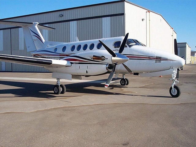 A white Beechcraft King Air with red and blue stripes is parked on an tarmac outside a hangar.