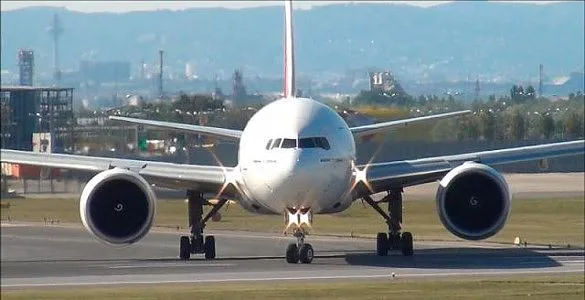 A large white twin-engine jet aircraft, likely a Boeing 777, is seen head-on on an airport taxiway with an urban background.