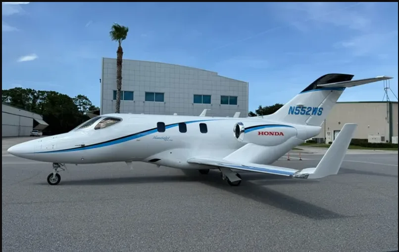 A side view of the white HondaJet HA-420, registration N552WS, with blue accents, parked on an airfield next to a hangar.