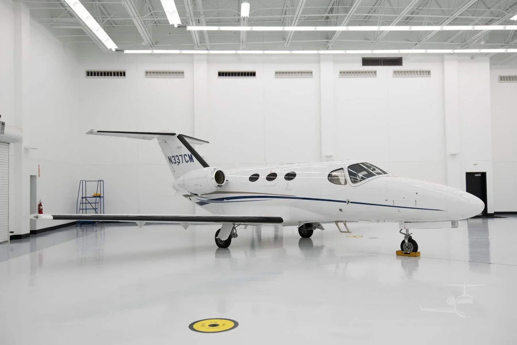 A white Cessna Citation M2 business jet with blue and gold stripes, tail number N337CM, parked in a brightly lit hangar.