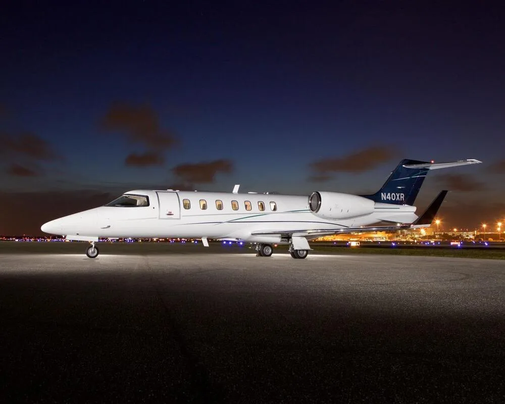 A white Learjet 40XR with a dark blue tail, parked on a tarmac at night with city lights in the background.