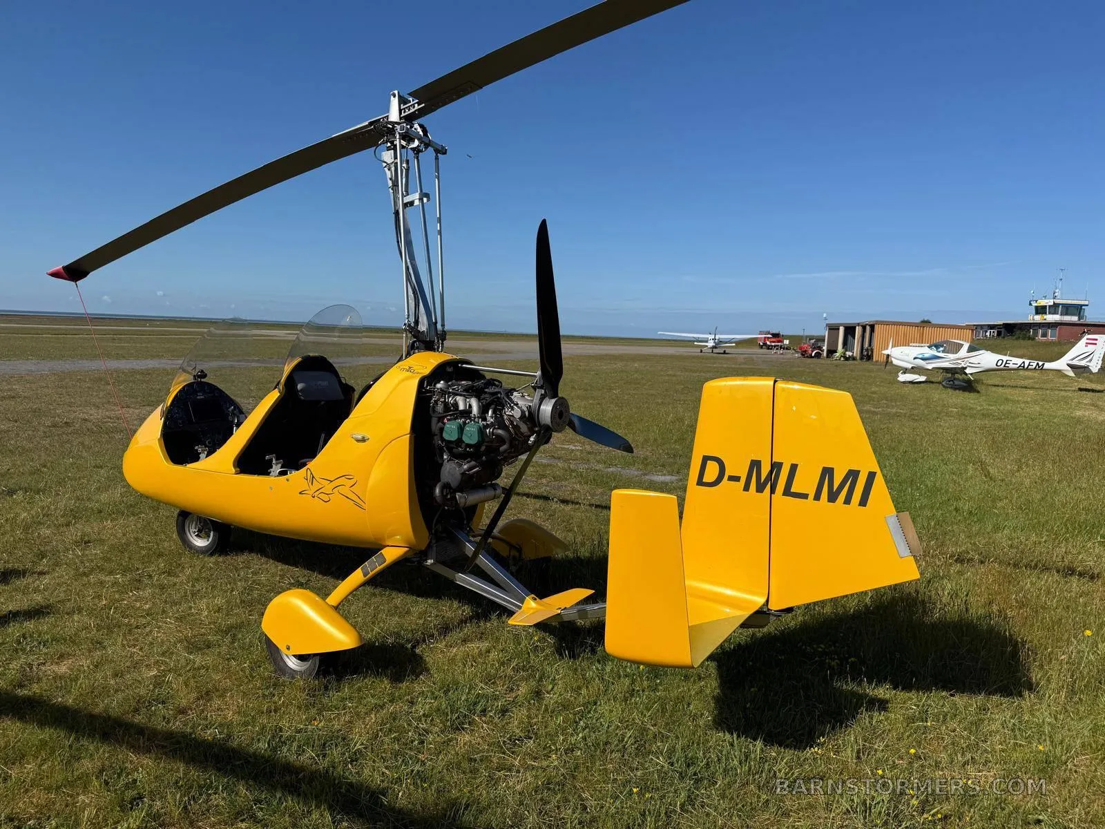A yellow MTO Sport autogyro (D-MLMI) with its exposed engine, parked on a grassy airfield with other aircraft in the background.
