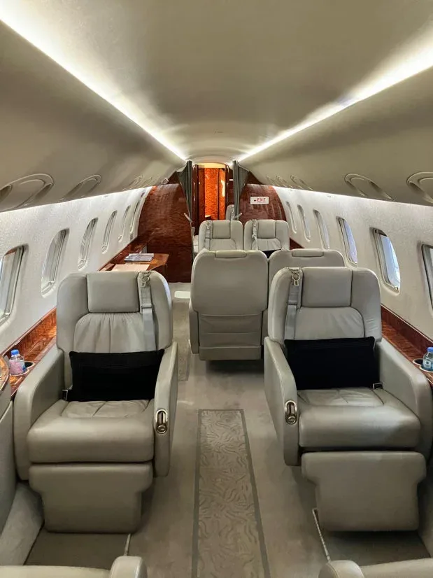 The interior cabin of a private jet features light grey leather seats, wood paneling, and overhead lighting.