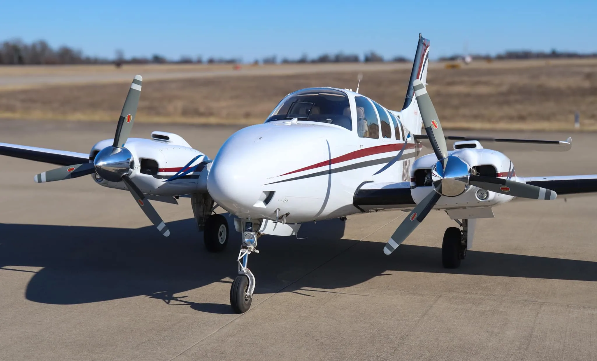 Front view of a white twin-engine Beechcraft Baron N204G with red, blue, and gray stripes, parked on an airfield.