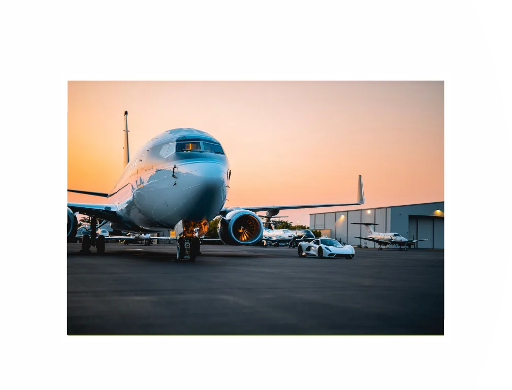 A Boeing 737 BBJ in a dark blue and white livery parked on an airfield at sunset, with a white sports car in front.