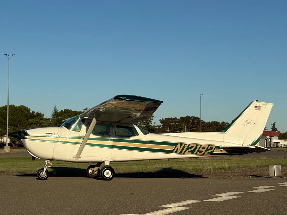 A white Cessna 172 with green and gold stripes parked on an airfield next to a grassy area.