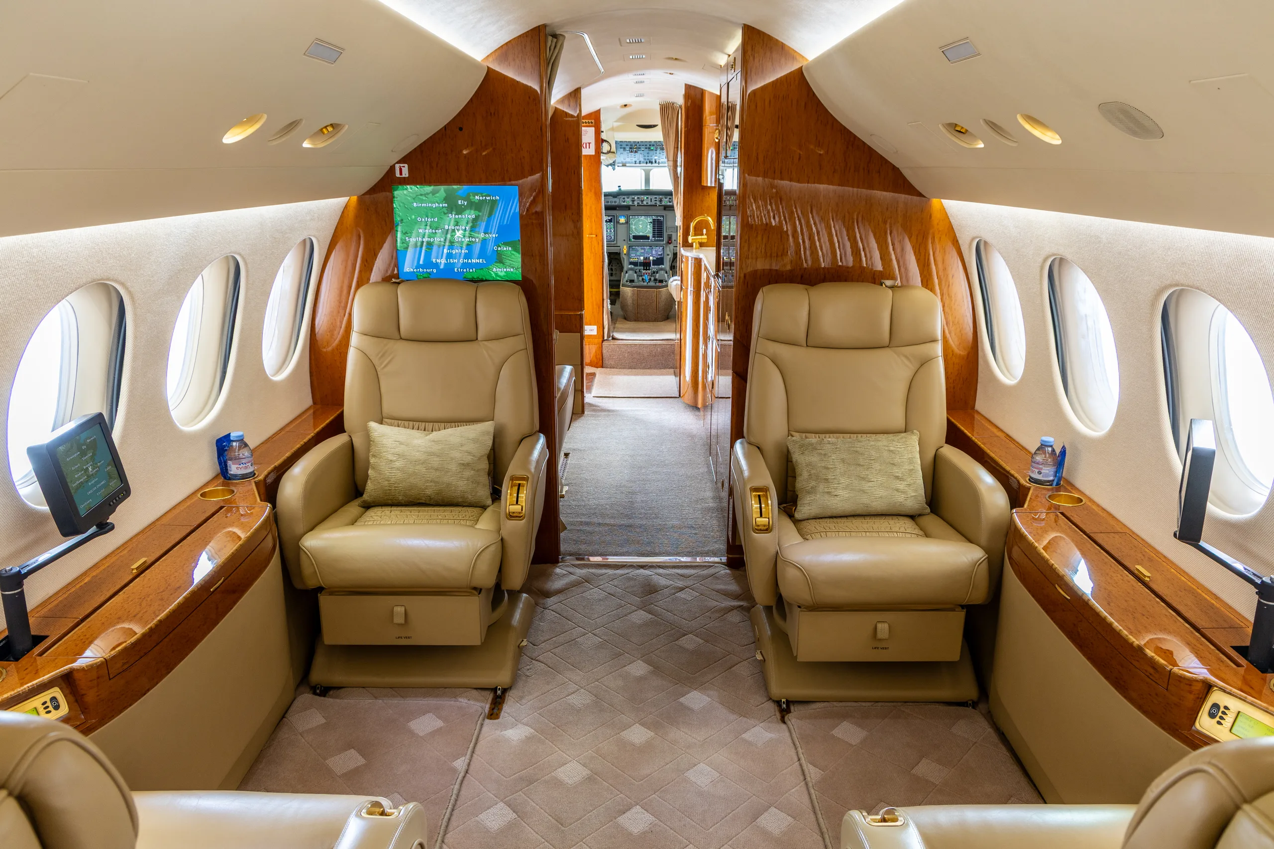 The luxurious interior cabin of a business jet with tan leather seats, wood paneling, and an entertainment screen.