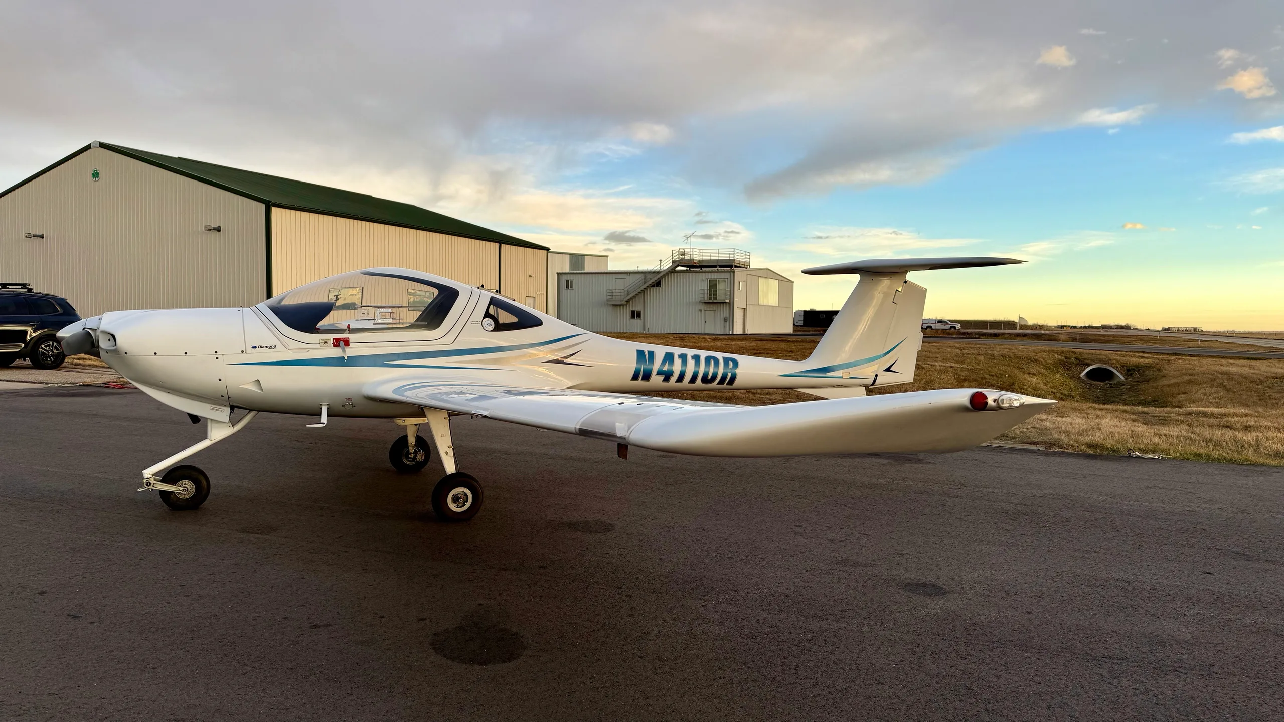 A white Diamond DA20-C1 Eclipse aircraft with blue stripes, registration N4110R, parked on an airfield next to a hangar.