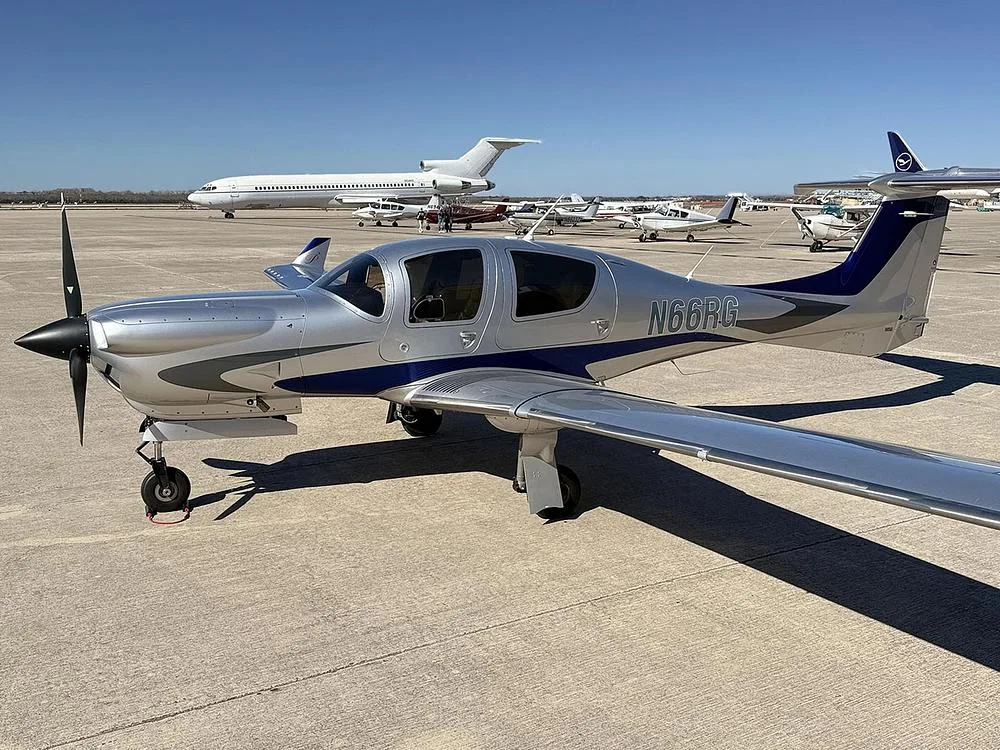 The silver and blue Diamond DA40 aircraft, N66RG, parked on an airfield with various other aircraft in the background under a clear sky.