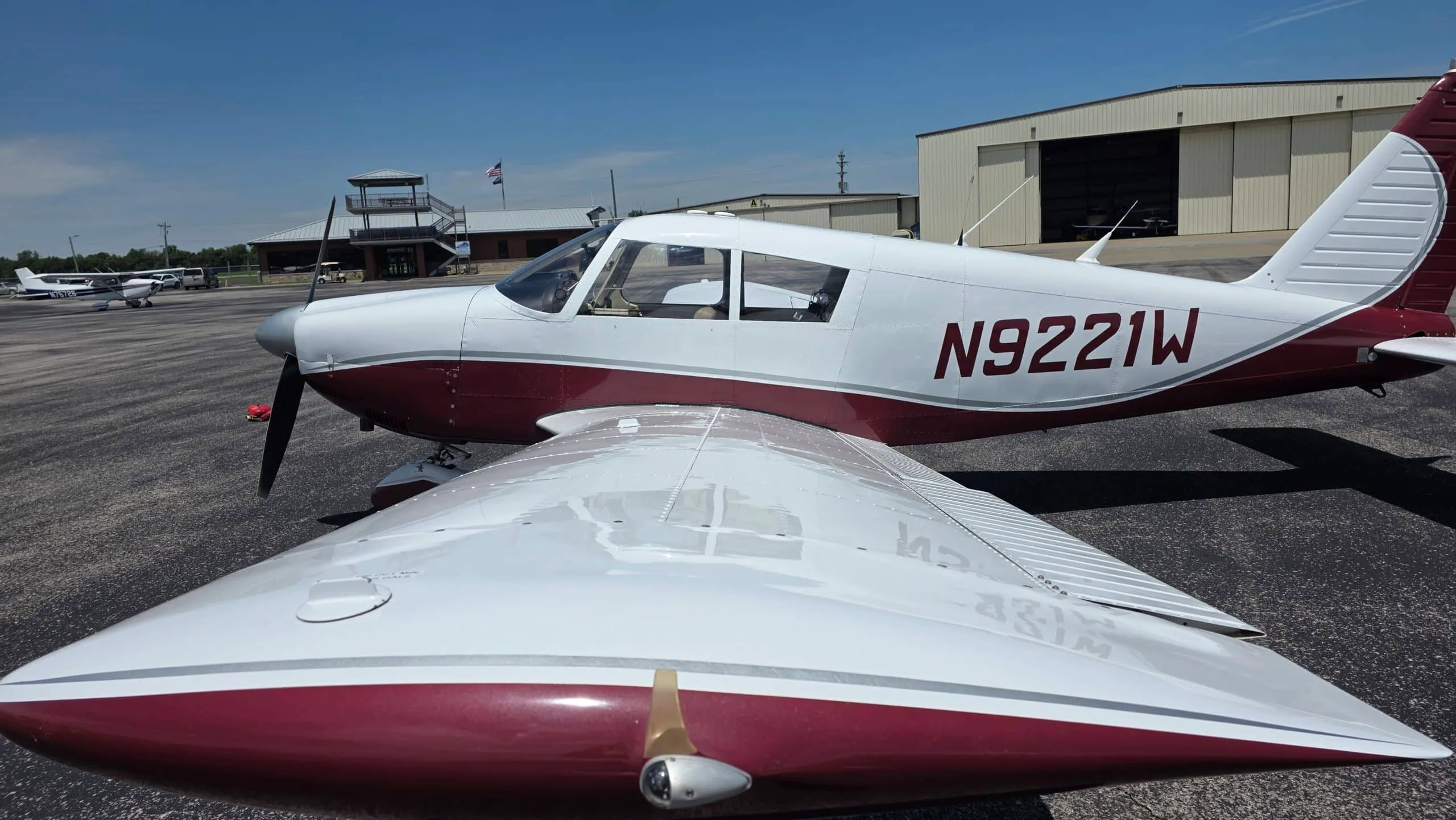 A view over the right wing of a white and maroon Piper Cherokee (N9221W) with a control tower in the background.