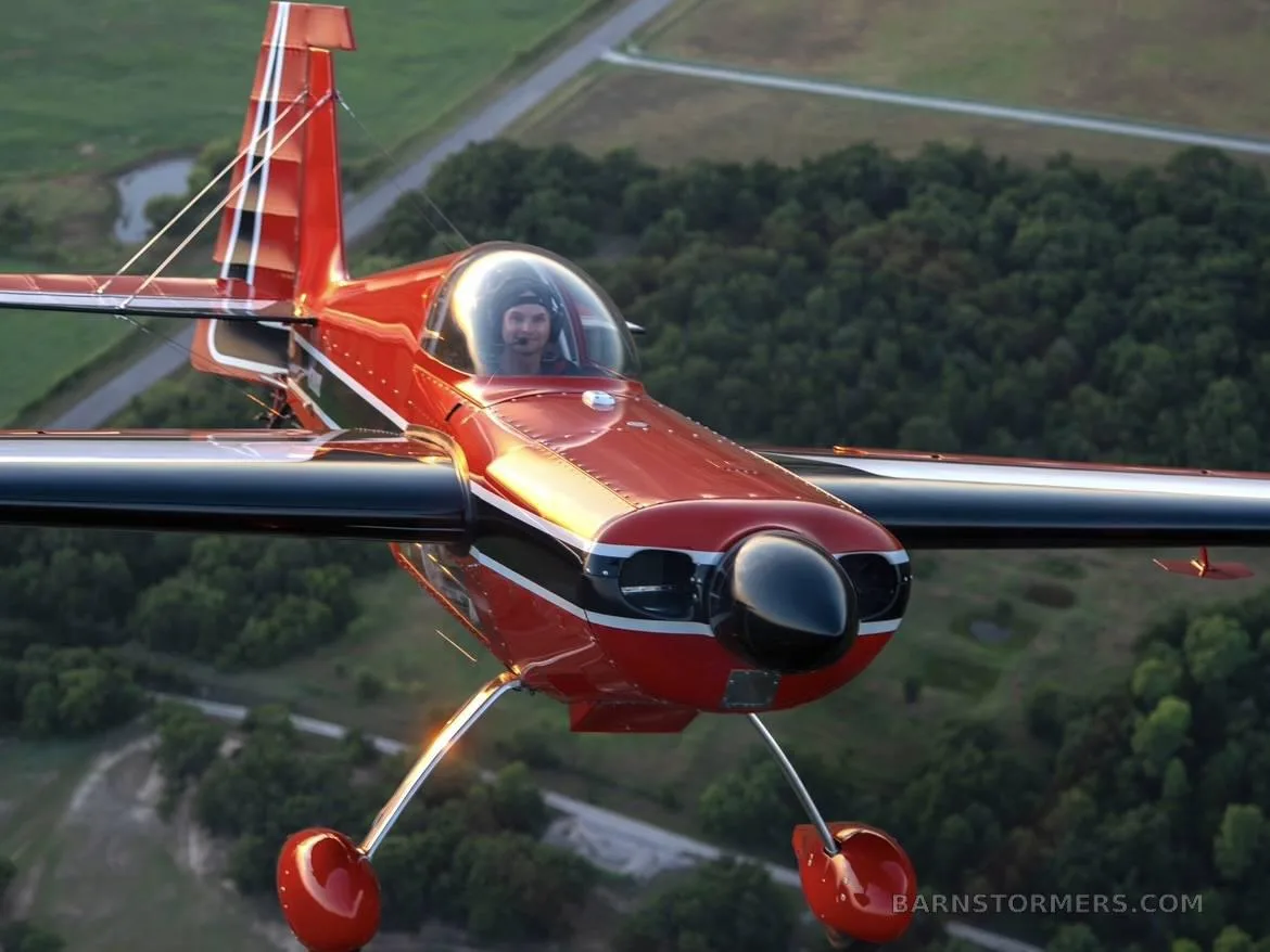 A close-up front view of a red and white aerobatic aircraft, N300CB, with a pilot, flying over green terrain.