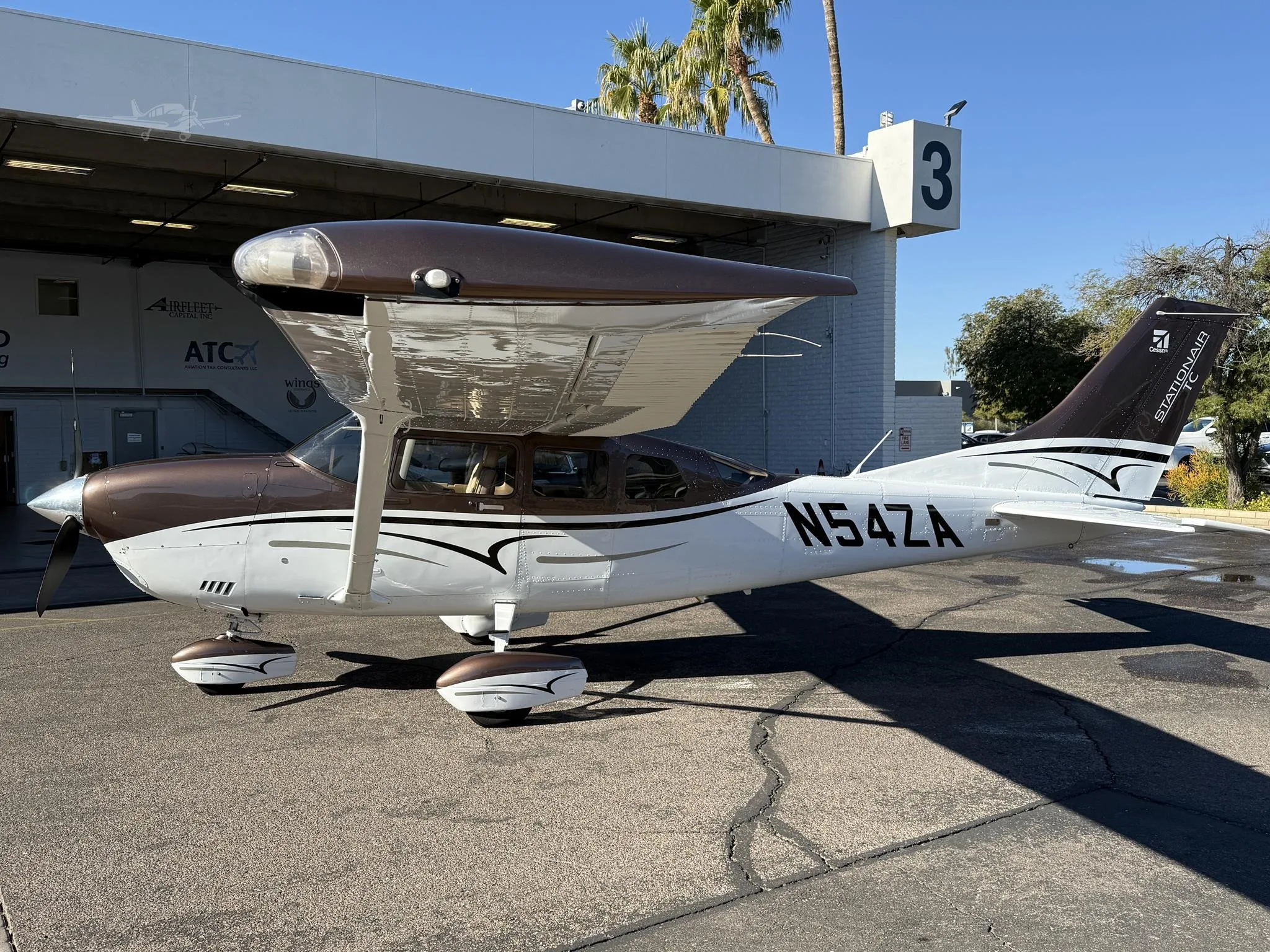 A white and brown Cessna Stationair TC (N54ZA) parked on an asphalt tarmac next to a hangar with palm trees in the background.