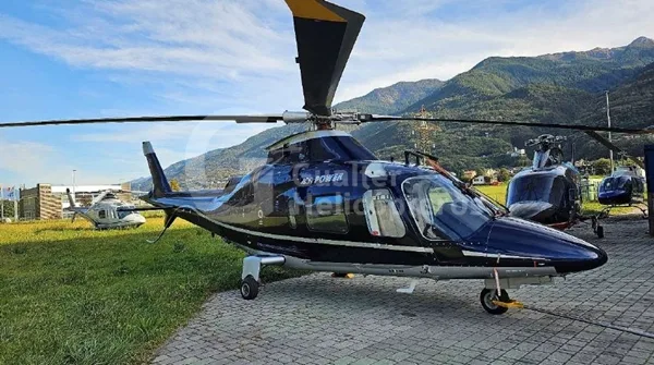 A dark blue Agusta A109 Power helicopter parked on grass next to a paved area with other helicopters and mountains in the background.
