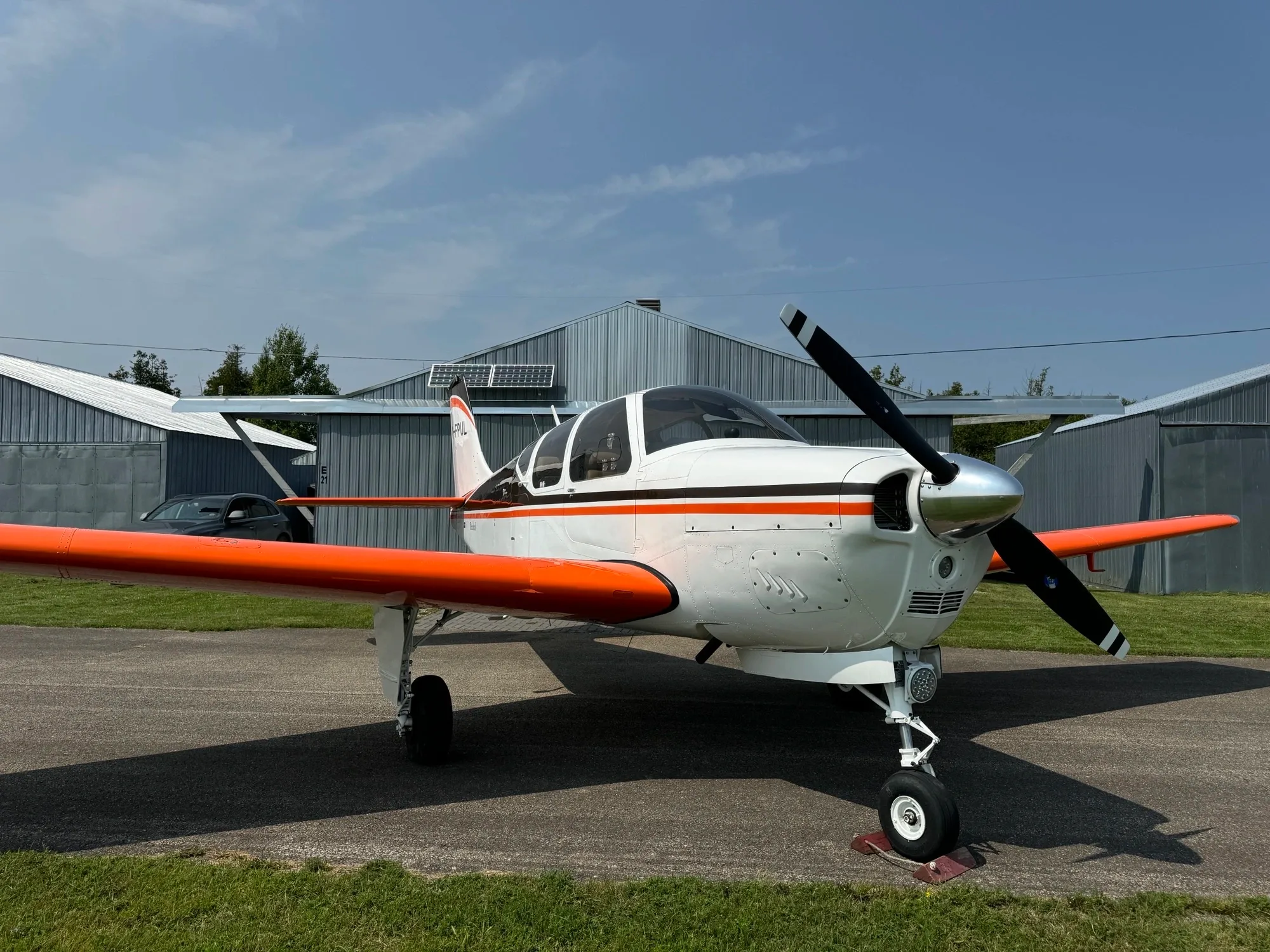 A white single-engine propeller aircraft with bright orange wings and tail is parked on an asphalt ramp in front of hangars under a clear sky.