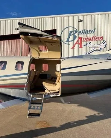 The side of a white and black aircraft, N850MR, with its main entry door open and stairs extended, parked in front of a hangar.
