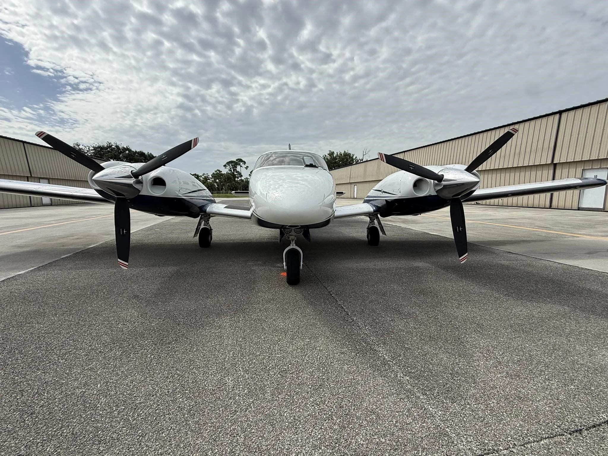 A head-on view of a white and blue Piper Seneca V aircraft parked on an airfield tarmac under a cloudy sky.