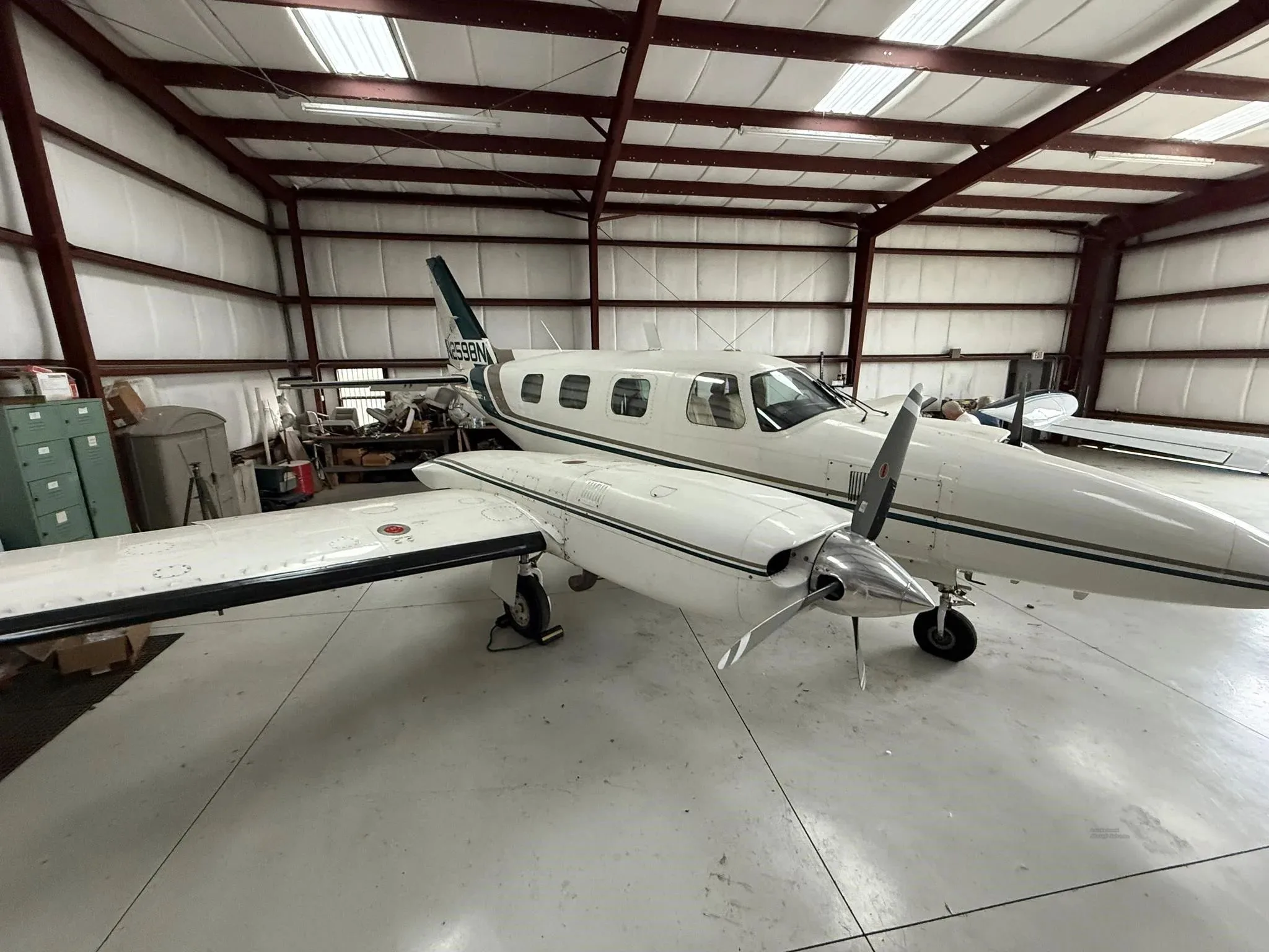 A white twin-engine Piper Navajo aircraft with green and brown stripes, seen from the front-right in a hangar.