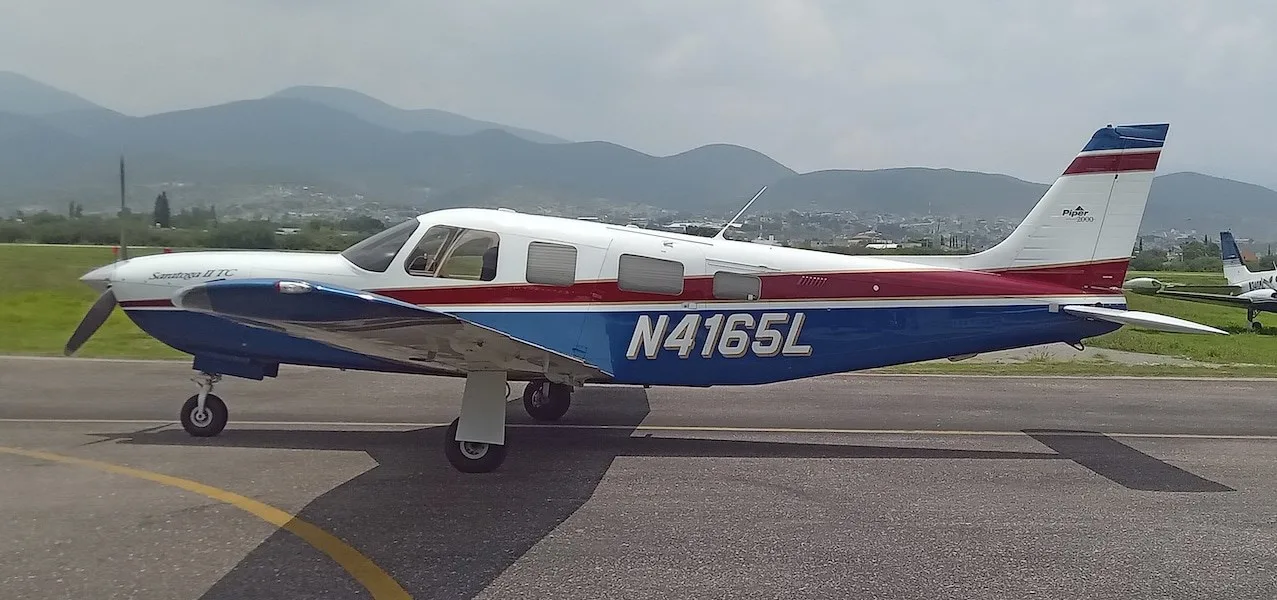 A Piper Saratoga II TC aircraft with white, blue, and red paint scheme on a tarmac with mountains in the background.