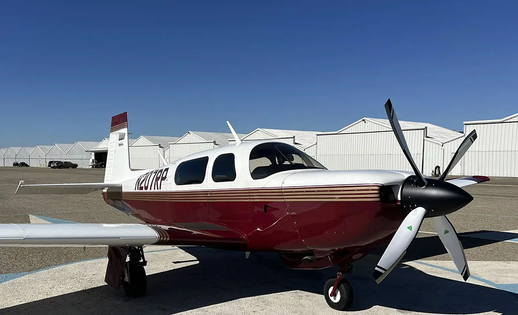 A white and maroon single-engine propeller aircraft, N207RP, is parked on an airfield with hangars in the background, viewed from the front-right.