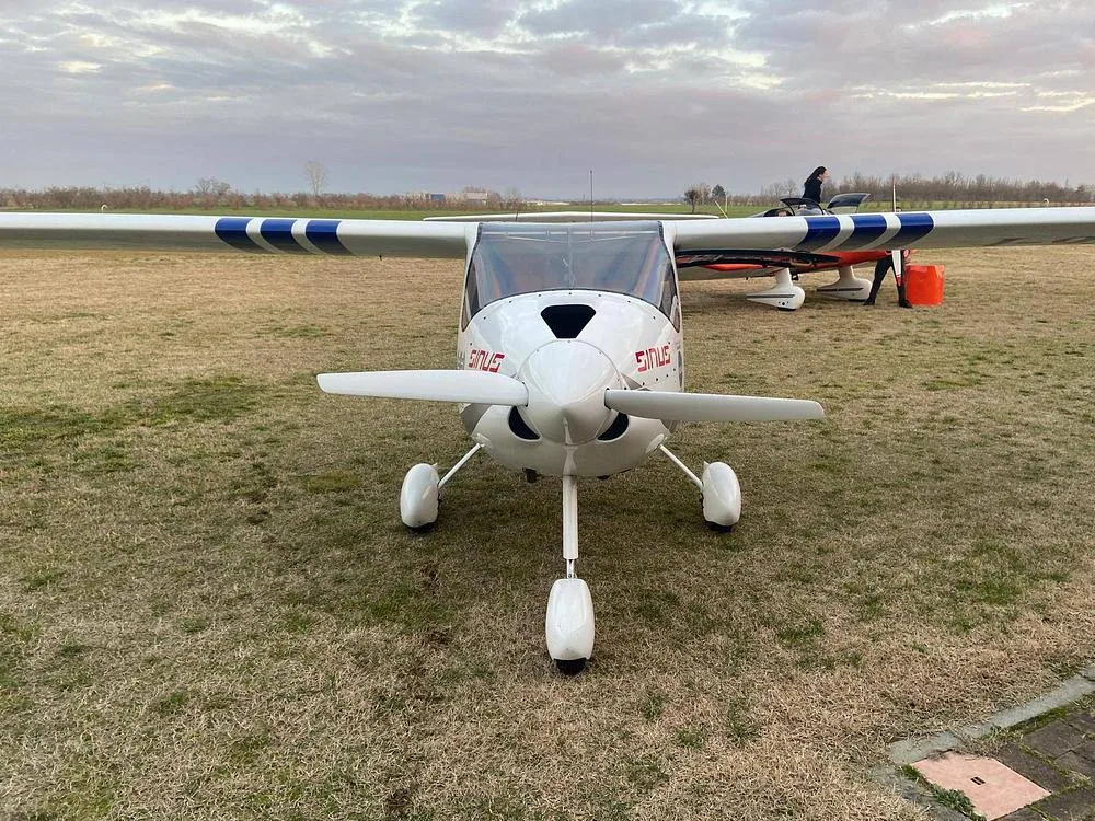 A white Pipistrel Sinus aircraft with blue wing stripes, viewed from the front on a grassy airfield under a cloudy sky.