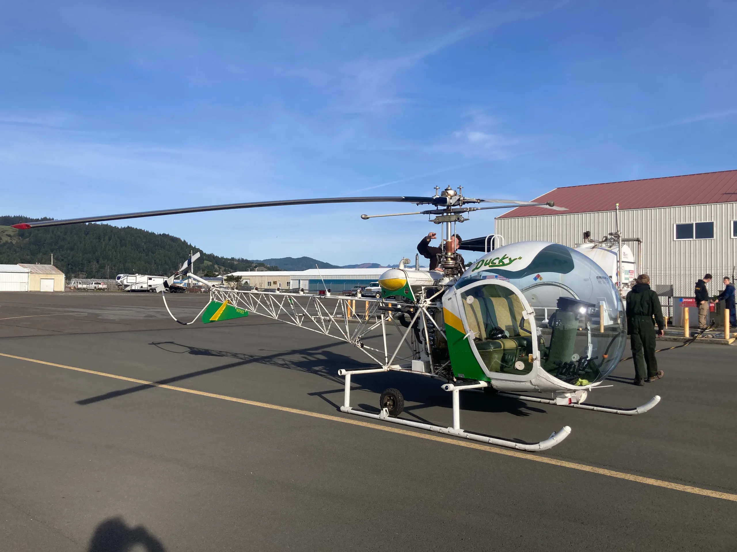 A white, green, and yellow open-frame helicopter named 'Ducky' sits on an asphalt tarmac with people, buildings, and hills in the background under a clear sky.