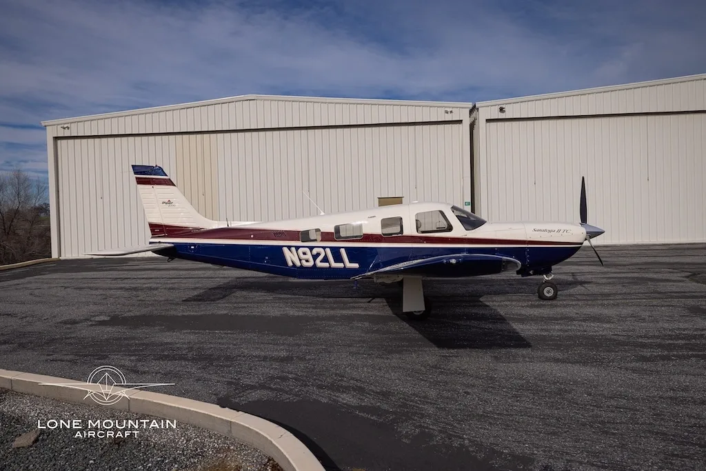 A side view of the Piper Saratoga II TC aircraft (N92LL) with blue and white paint scheme parked on an asphalt apron in front of hangars.