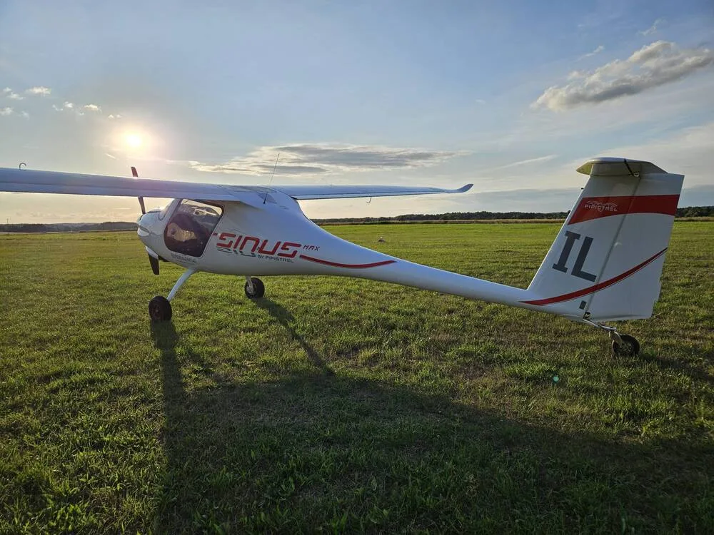 A white Pipistrel Sinus aircraft with red accents and "IL" tail number is parked on a grassy airfield at sunset.