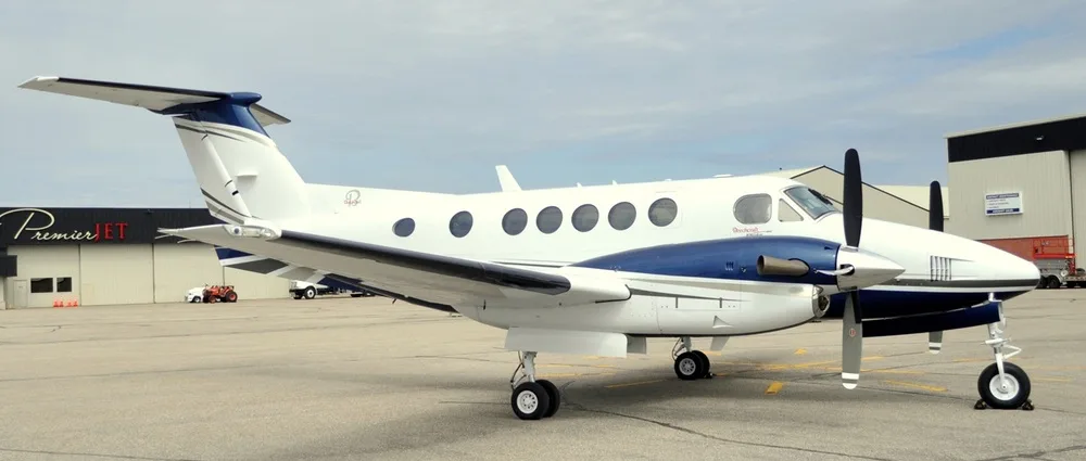 A white and blue twin-engine turboprop aircraft, likely a Beechcraft King Air, parked on an tarmac in front of a hangar.