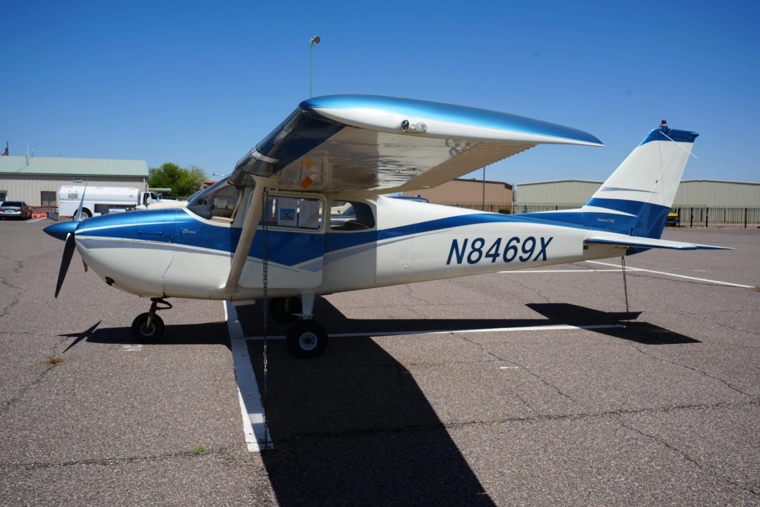 Side view of a white and blue Cessna 172 (N8469X) with its tail facing right, parked on an airfield.