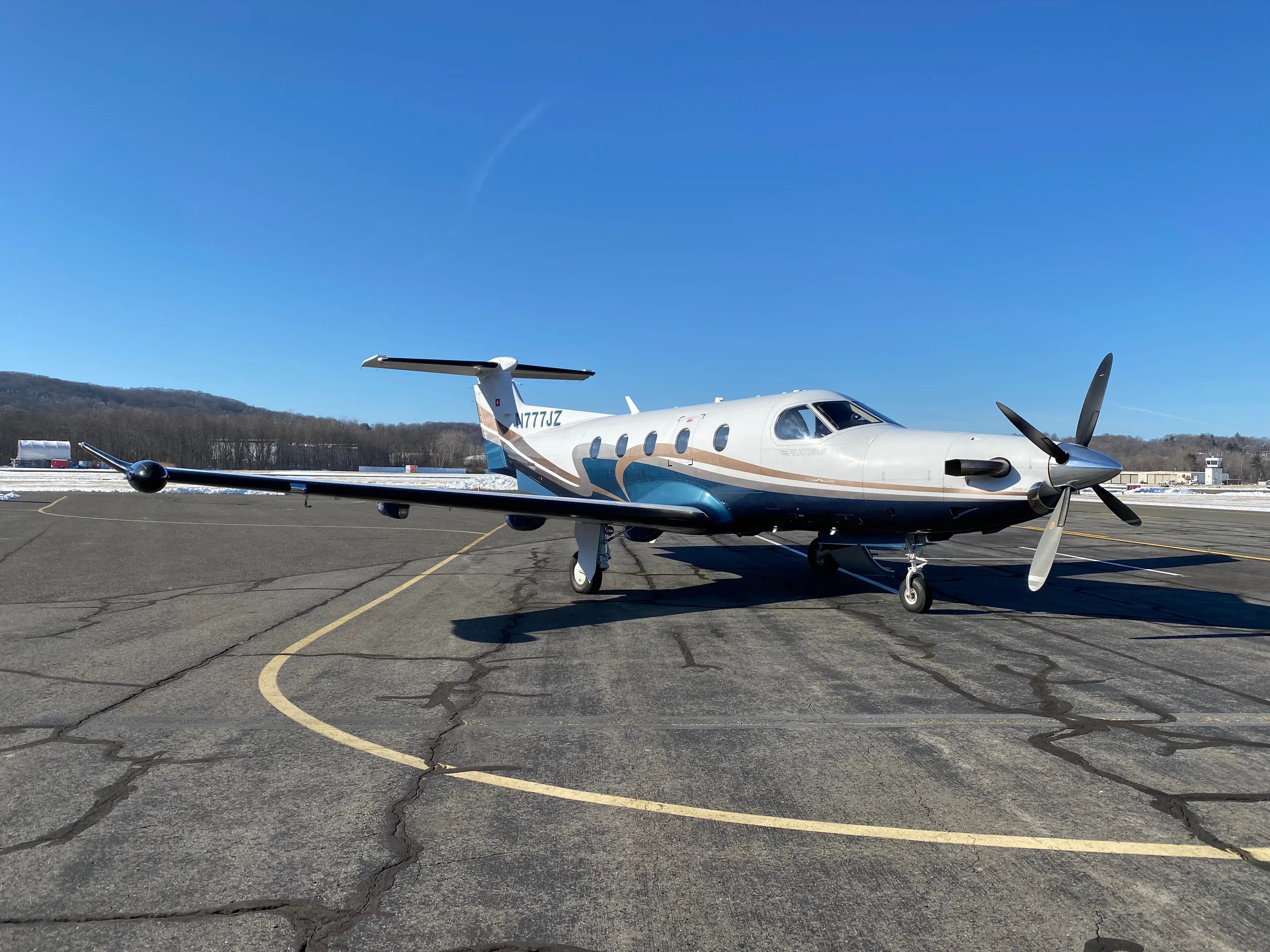 A Pilatus PC-12 aircraft with registration N777JZ is parked on the tarmac with a blue and brown paint scheme.