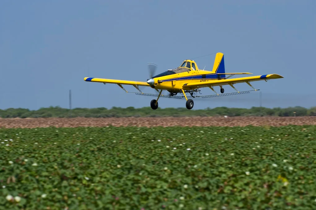 A yellow and blue agricultural aircraft, N1740A, flies low over a green field with white blooms.