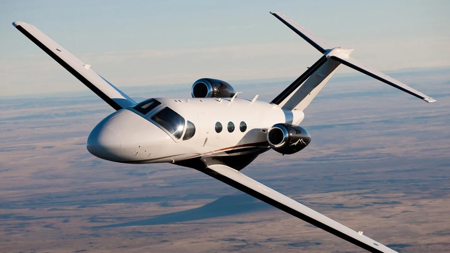 A white business jet with black and brown accents and a mountain logo on its engines flies over a desert landscape.