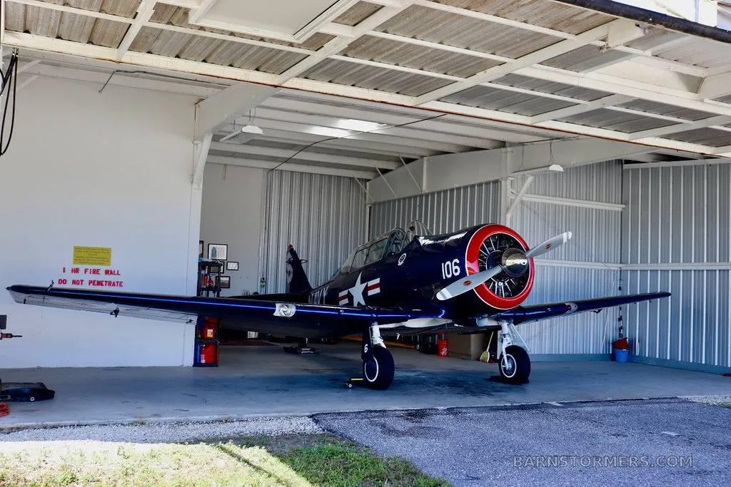 A dark blue vintage military trainer aircraft with a red nose is parked inside a white hangar.