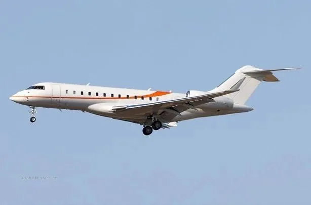 A white private jet with orange stripes on its fuselage and landing gear extended, flying against a clear blue sky.