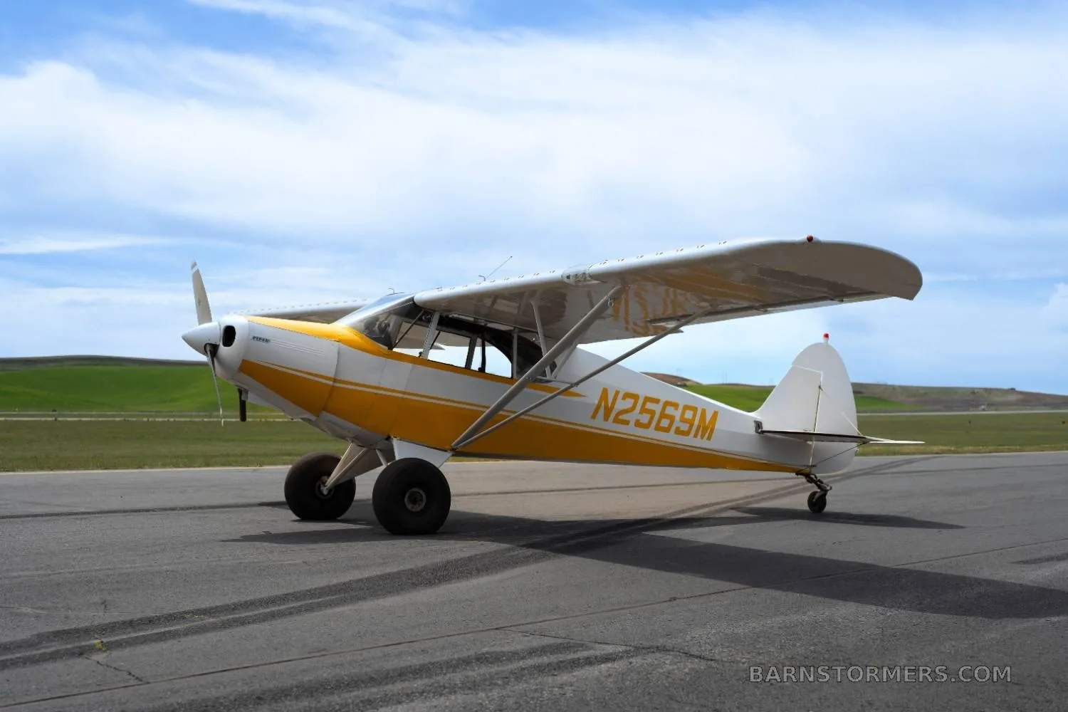 Side view of a yellow and white single-engine taildragger aircraft (N2569M) with large tires on an asphalt runway.