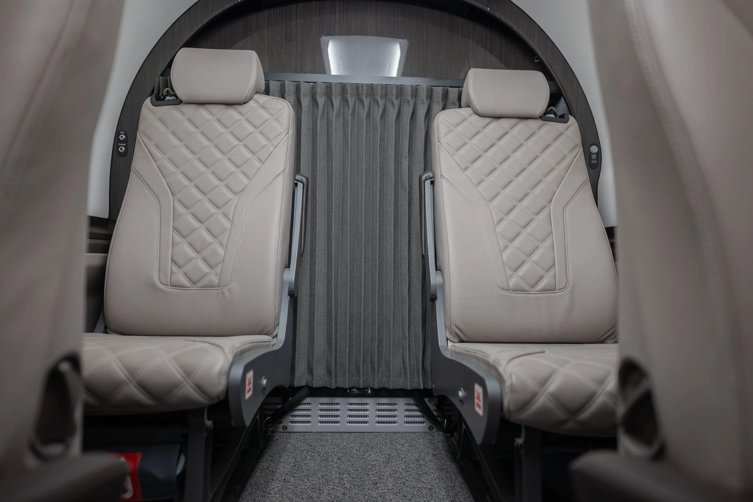 Two light grey quilted leather seats in the cabin of a Pilatus PC-12 aircraft, with a privacy curtain in the background.