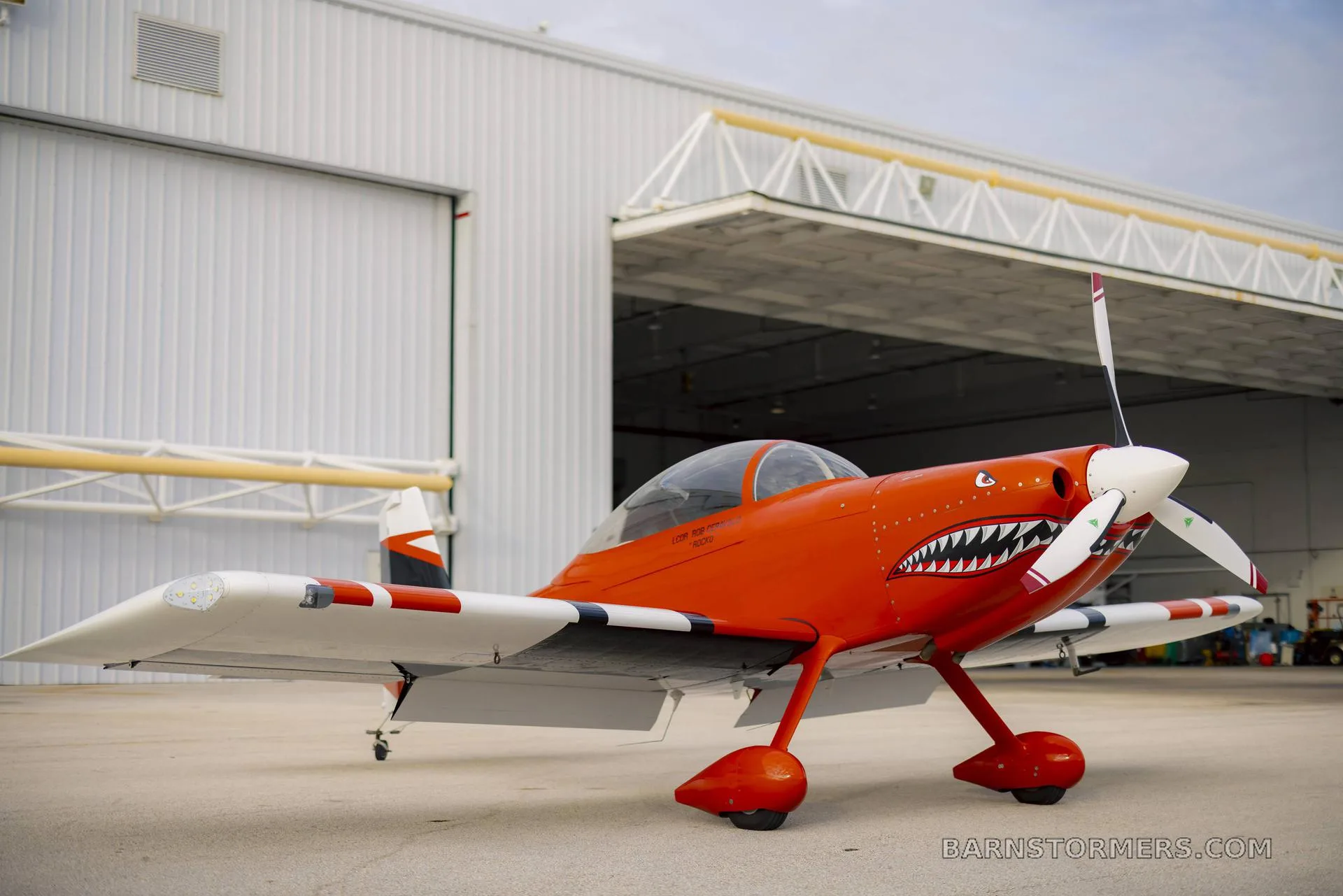 A low-angle front-side view of an orange and white single-engine aircraft with a shark mouth paint scheme, parked on an airfield.