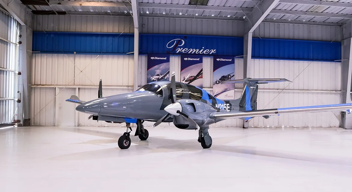 A gray and blue Diamond DA62 aircraft, registration N215E, parked inside a hangar.