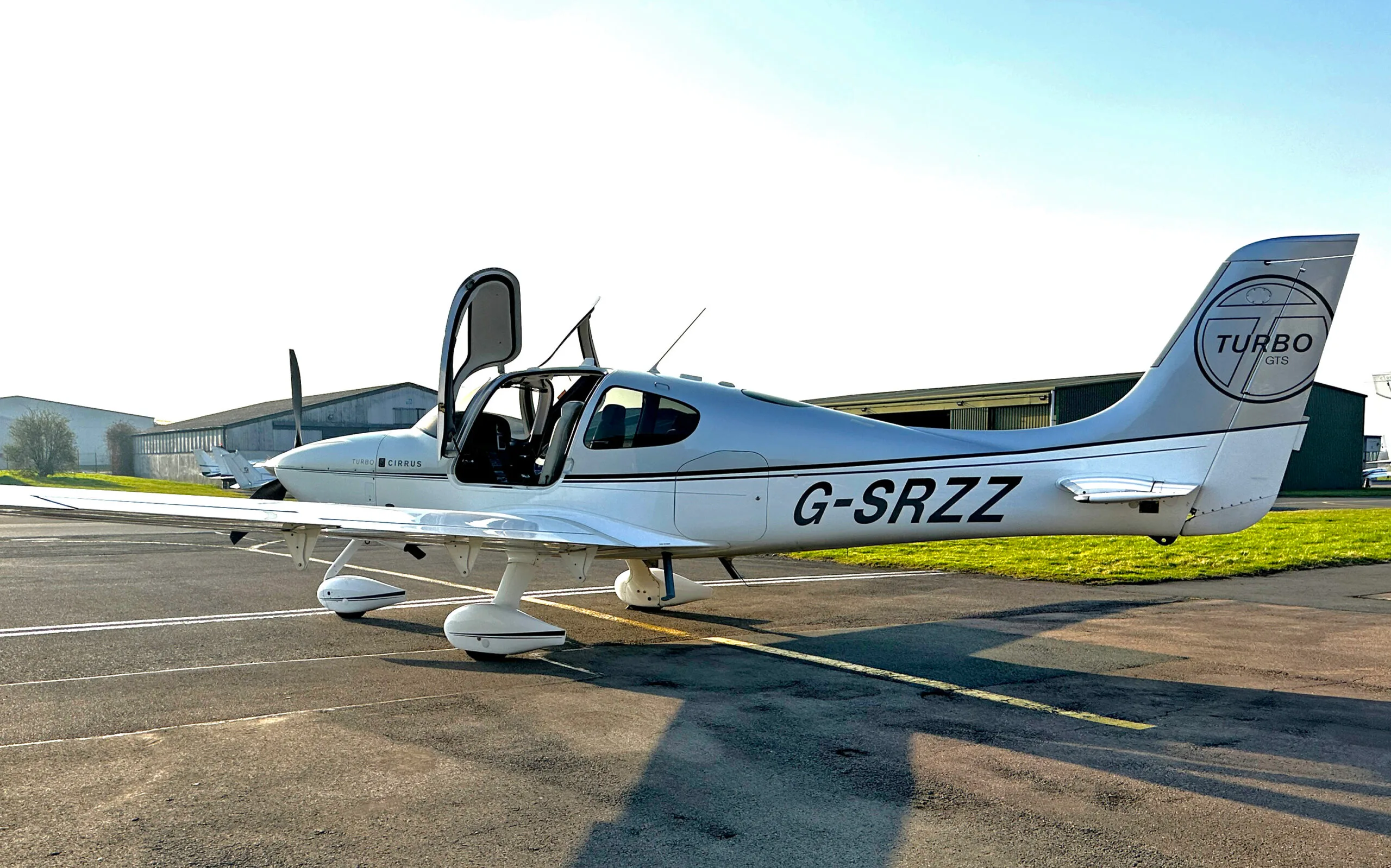 A silver and white Cirrus SR22 Turbo aircraft with registration G-SRZZ and an open door, parked on an airfield.