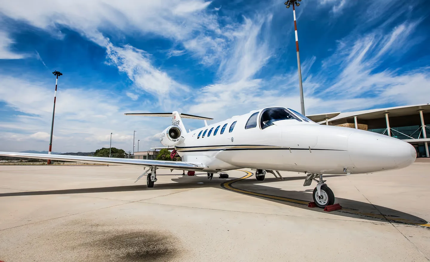 A white business jet with a gold stripe is parked on an airport tarmac under a blue sky with wispy clouds, with an airport building in the background.