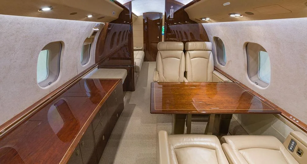 The elegant cabin interior of a business jet, showcasing a polished wooden table and beige leather seating.