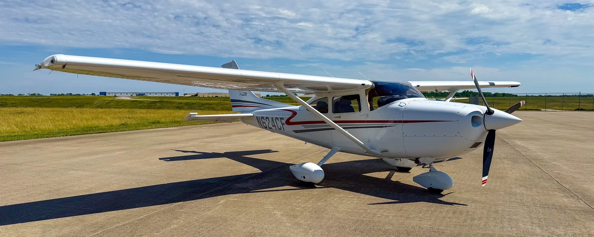 A white Cessna 182 Skylane with red and gray stripes, registration N624CF, parked on an asphalt ramp with a grassy field in the background.