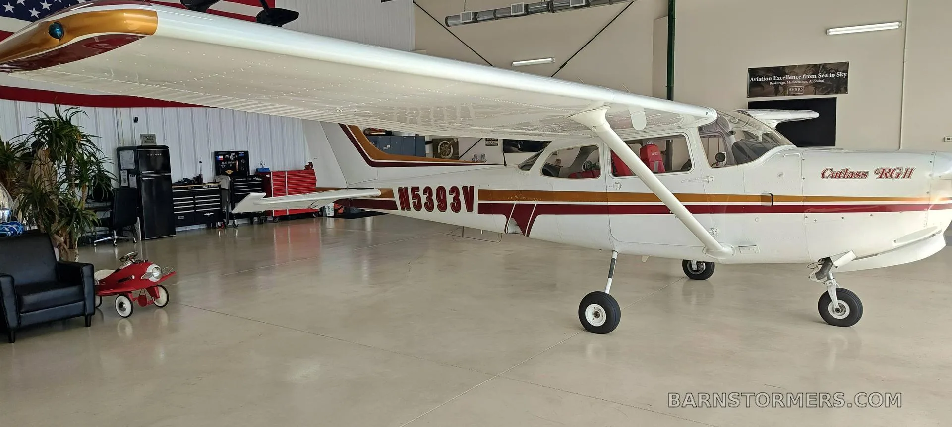 A white Cessna Cutlass RGII (N5393V) with red and gold stripes is viewed from the left side inside a hangar.