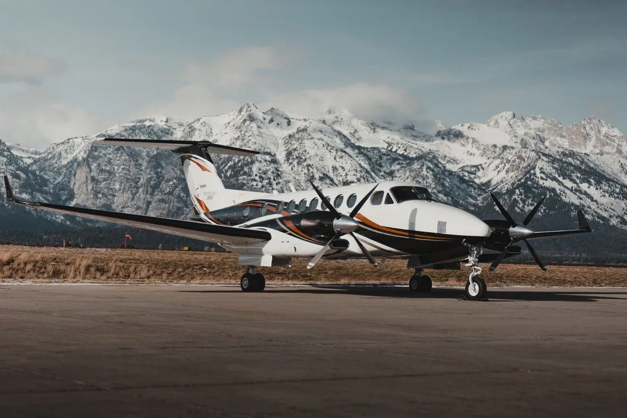 A white, black, and orange King Air 350i aircraft parked on an airfield with snow-capped mountains in the background.